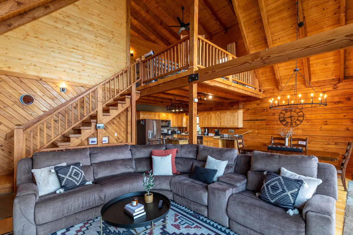 A spacious living area with a large sectional sofa facing an open kitchen area and a wooden staircase. The room is warmly lit, with a chandelier hanging from the ceiling, and offers a view of the kitchen, perfect for group activities and gatherings.