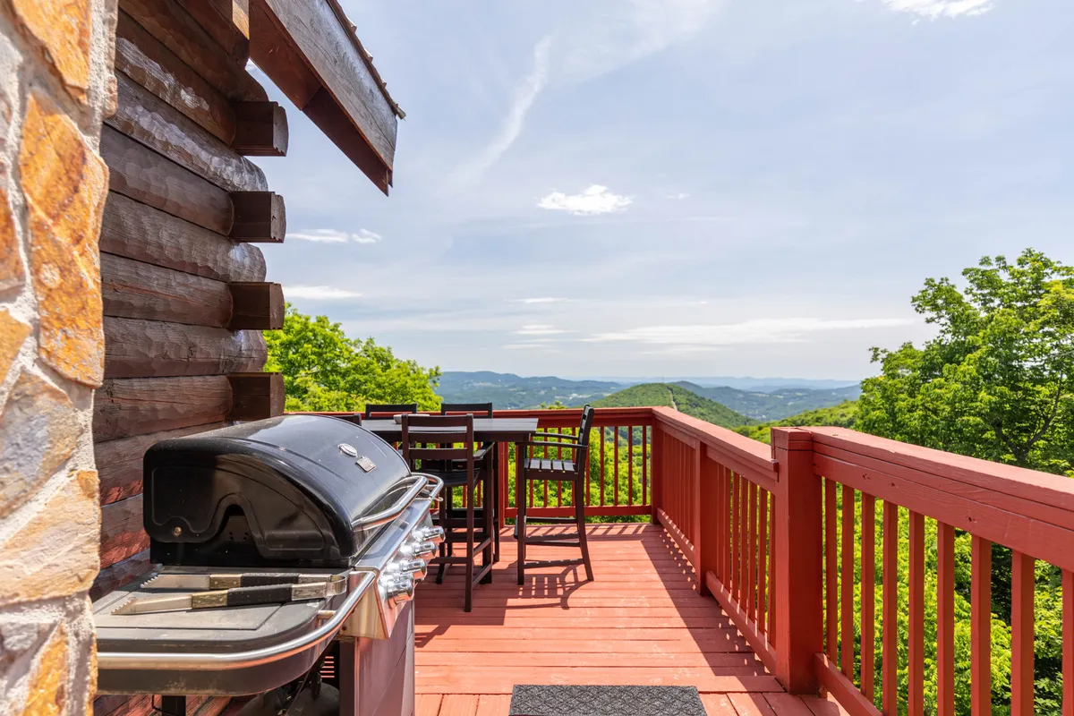 A wooden deck with a dining set and grill overlooks sweeping mountain vistas. The scene invites guests to dine outdoors while enjoying the natural beauty surrounding the cabin.
