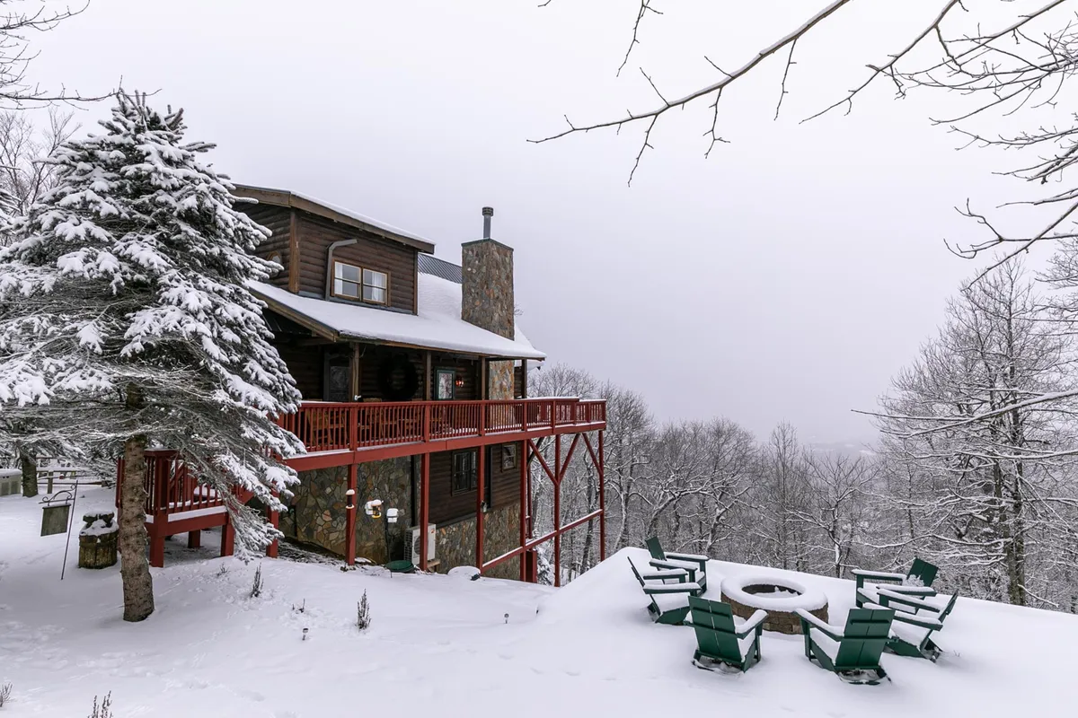 A snow-covered mountain landscape surrounds a wooden deck with a fire pit. The scene captures the inviting outdoor space with Adirondack chairs around the fire, framed by snow-laden trees and panoramic mountain views.