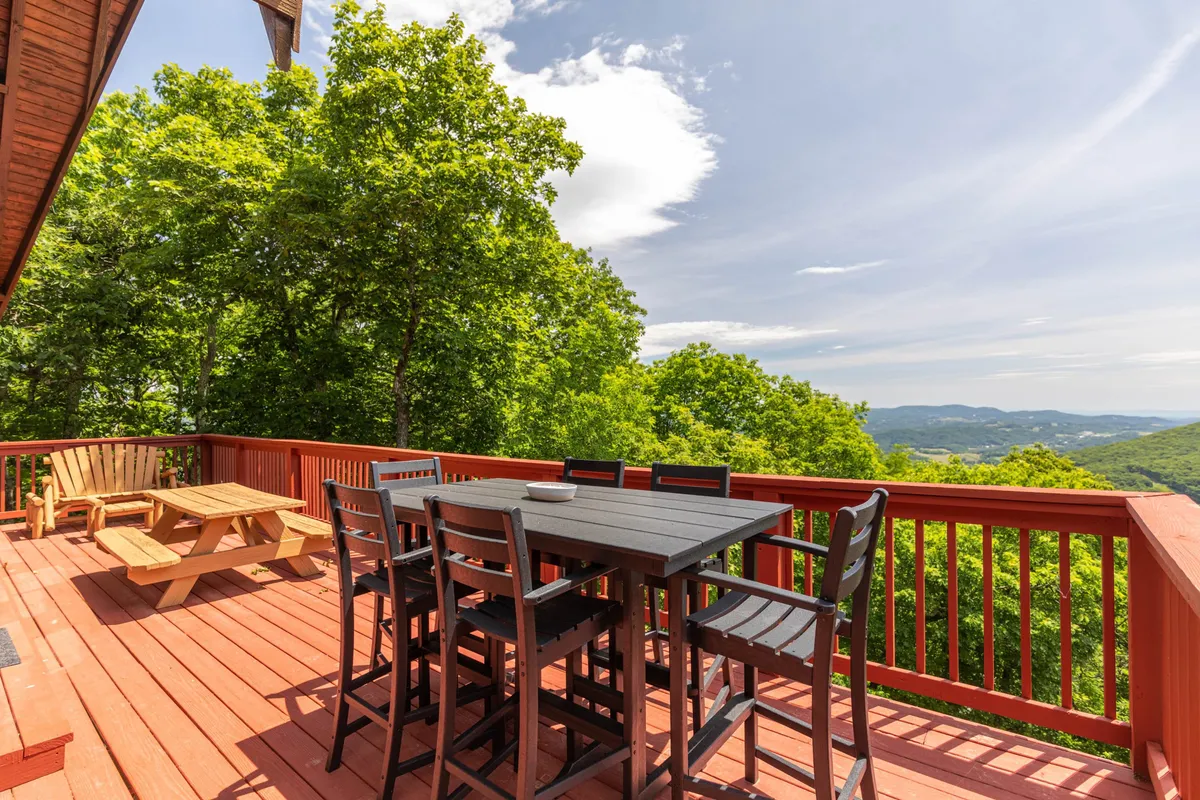 A wooden deck furnished with a dining table and high chairs overlooks a lush green landscape and distant mountain peaks. It’s an inviting setting for meals, morning coffee, or simply enjoying the fresh air and views.