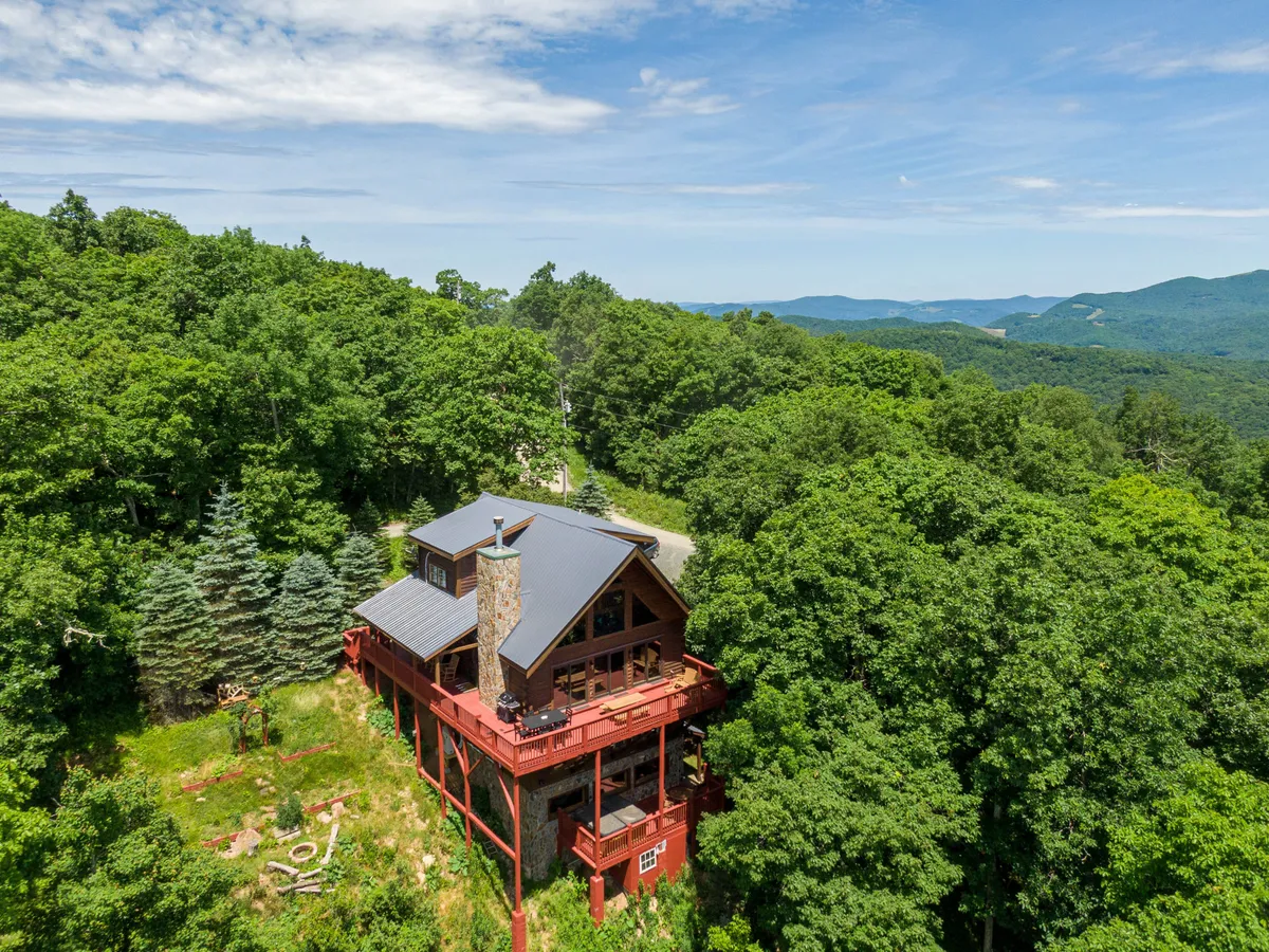 The hero image showcases a charming mountainside cabin perched on sturdy stilts, framed by lush trees and a striking stone chimney. The expansive deck offers breathtaking, unobstructed views of rolling Blue Ridge mountains stretching into the distance, ideal for soaking in sunsets or stargazing. The tranquil setting highlights the cabin's seclusion and connection to nature, with plenty of outdoor space for relaxing, dining, or exploring the surrounding woods.