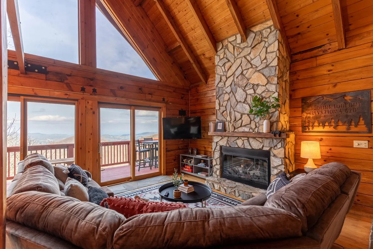 This interior image highlights a large stone fireplace, spacious couch, large windows, and a coffee table. The space is arranged for relaxation, watching TV, and gathering for group activities.