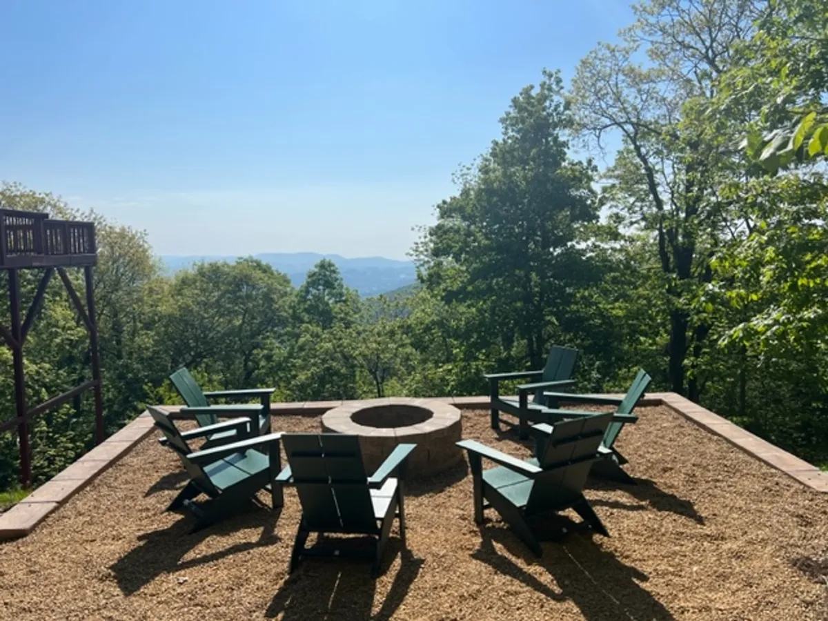 The scene centers on a rustic fire pit encircled by adirondack chairs, set against a sweeping mountain backdrop and lush trees. The area invites communal gatherings, storytelling, and enjoying the warmth of a crackling fire under open skies.
