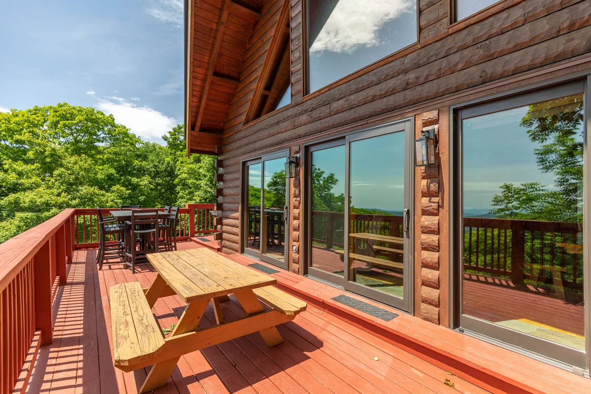 This image captures an inviting wooden picnic table on a spacious deck, set against lush greenery and large sliding glass doors leading inside. The scene suggests ample outdoor space for dining, relaxing, and play, with natural surroundings perfect for both people and pets.