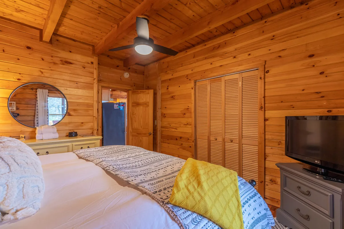 A cozy king bedroom with wooden walls and ceiling, a plush bed adorned with a decorative throw, a mirror, a dresser, and a wall-mounted TV. Soft natural light filters through, creating an inviting and relaxing atmosphere perfect for restful nights.