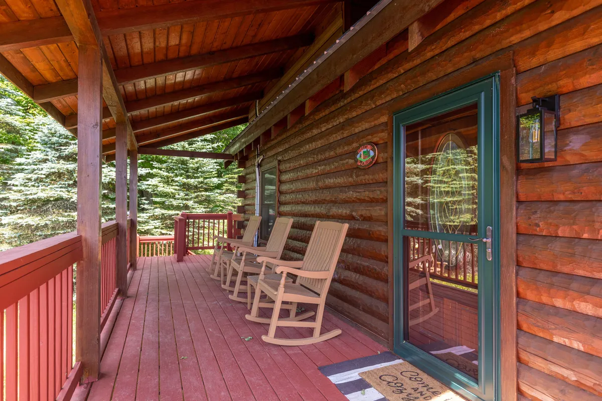 A peaceful log cabin porch featuring inviting wooden rocking chairs and vibrant red railings. The porch overlooks wooded surroundings, creating a cozy outdoor nook perfect for couples or friends to relax, watch sunsets, or stargaze in privacy.
