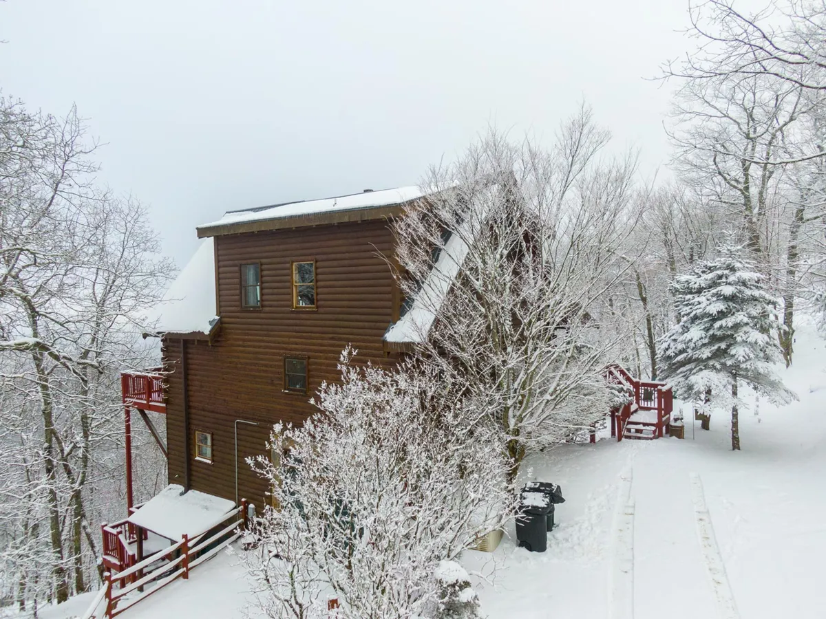 A wooden house sits surrounded by a snowy landscape—snow-covered trees, a red deck, and a pathway, all blanketed in winter white. The image conveys both the beauty and adventure of winter in the mountains.